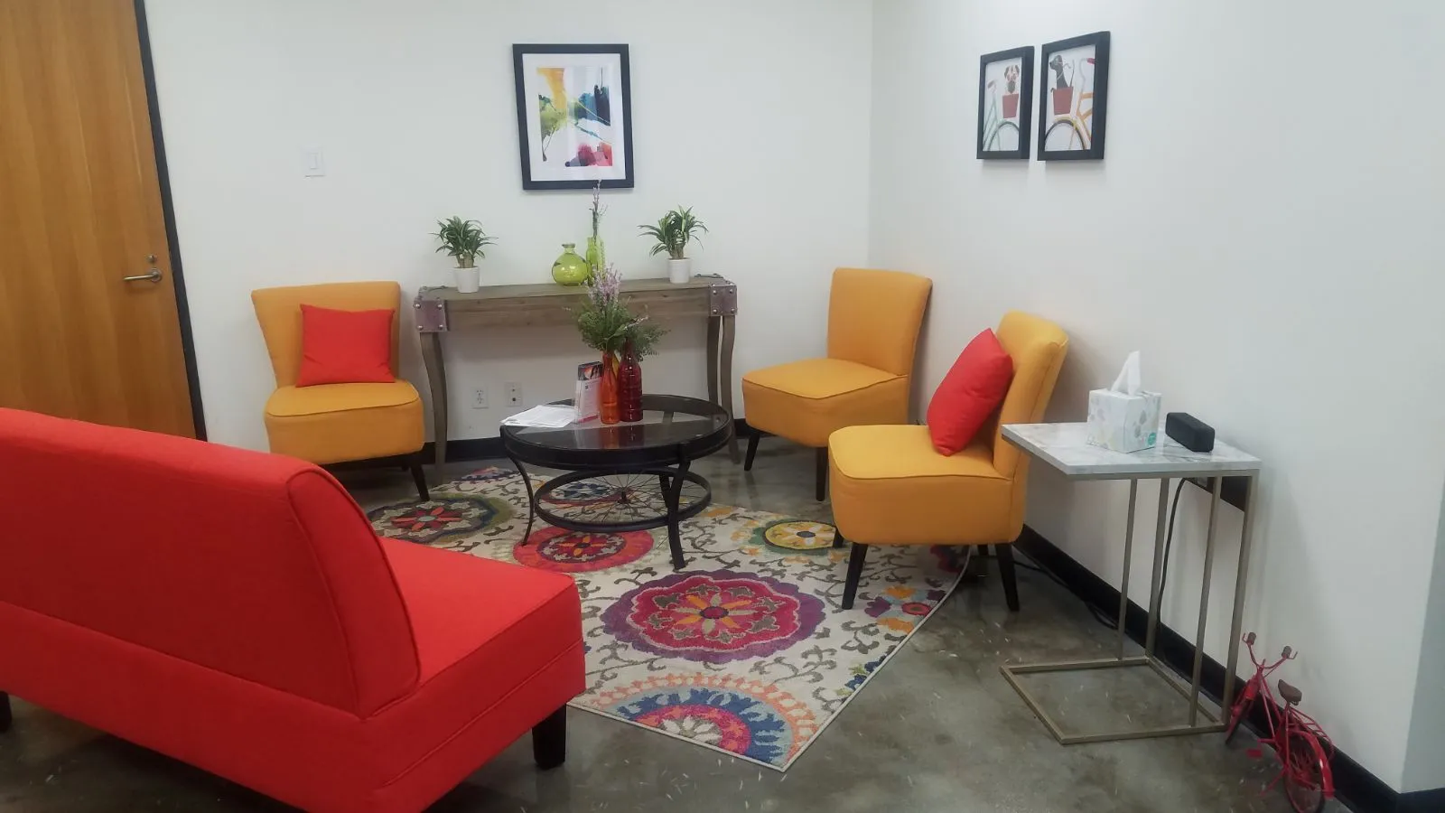 TMS therapy clinic treatment room with red and yellow chairs, black table, framed artwork, and side table with supplies