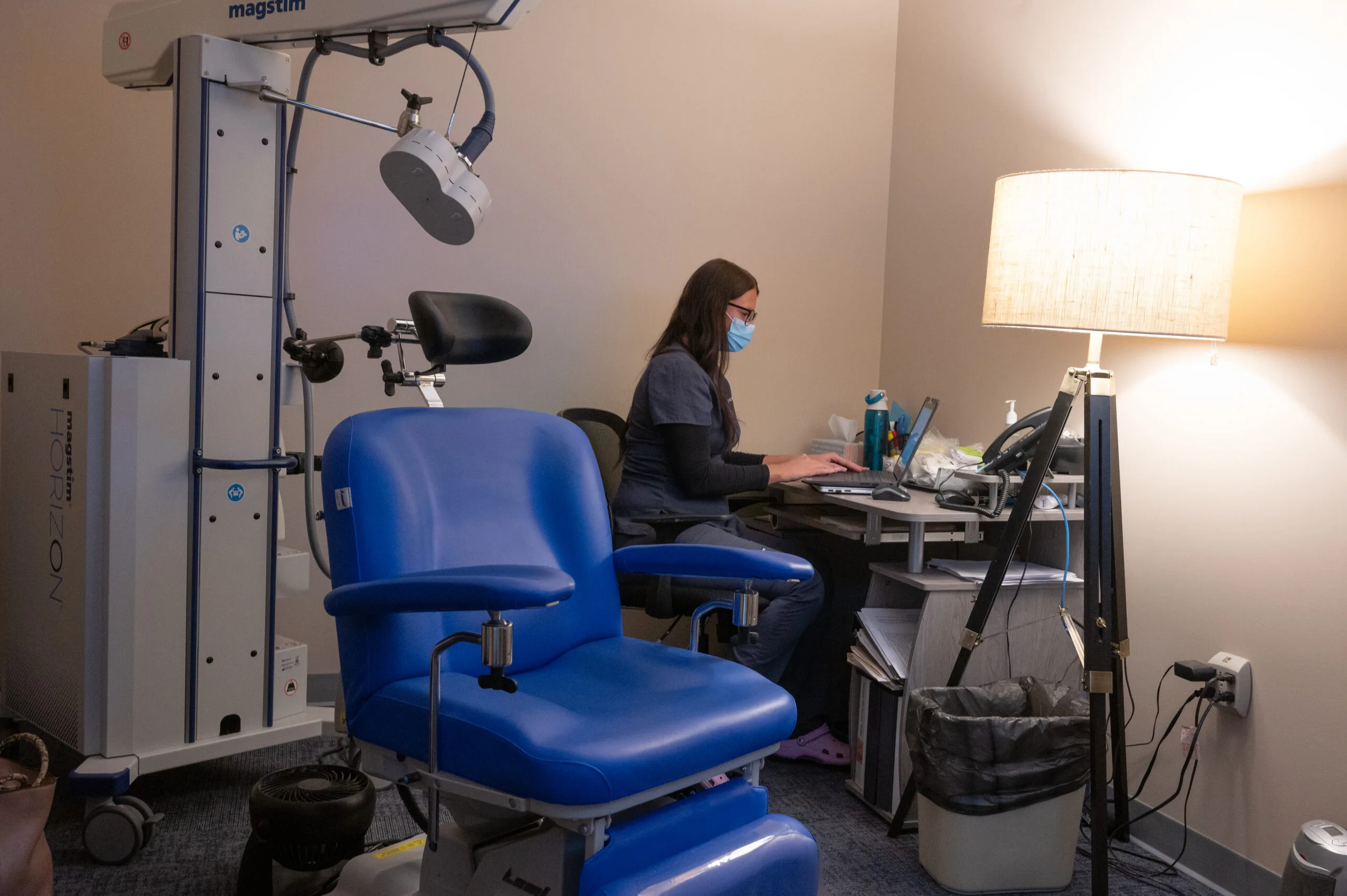 Staff member seated at desk beside blue TMS treatment chair with equipment overhead