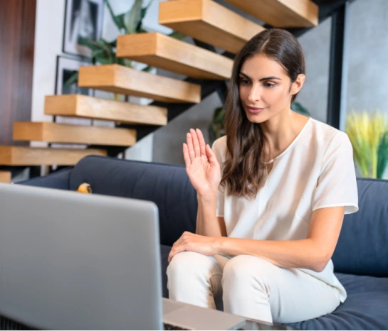 Woman waving at laptop during virtual telehealth session in modern living room