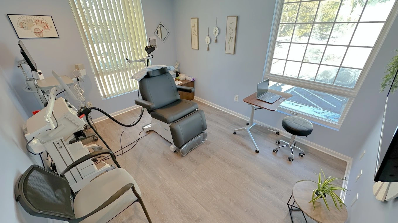 TMS treatment room with clinical chair, coil apparatus overhead, desk with monitor, and large windows with natural light