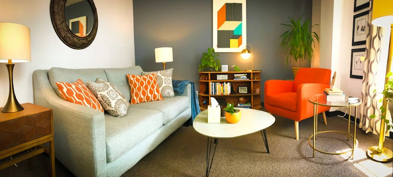 Therapy office waiting room with gray sofa, orange accent chairs, bookshelf, and plants