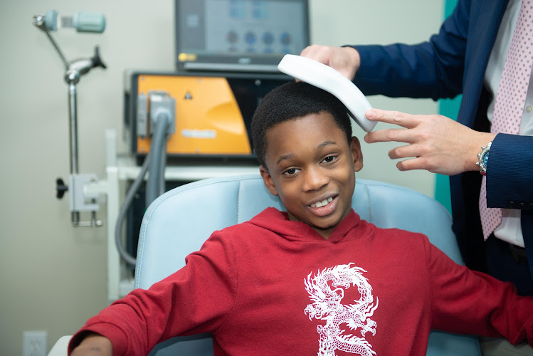 Clinician applying TMS treatment headpiece to smiling child in treatment room with equipment visible