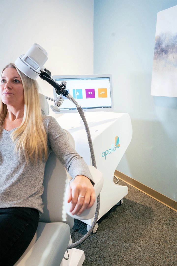 Patient receiving Apollo TMS treatment with coil positioned on head, monitor displaying session in background