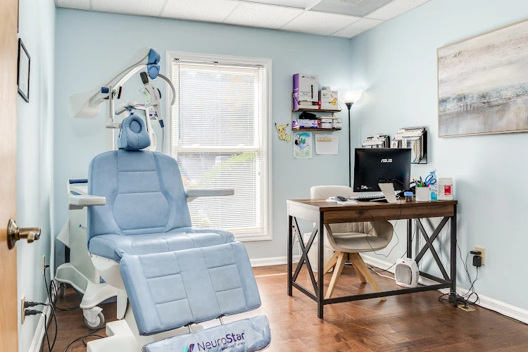 TMS therapy treatment room with blue padded chair, overhead magnetic coil device, desk with computer, and NeuroStar equipment