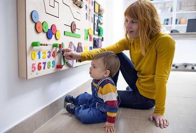 Therapist in yellow sweater guiding young child in interactive learning activity with colorful educational wall board