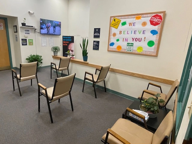 Outpatient clinic waiting room with chairs, plants, bulletin board reading 'WE BELIEVE IN YOU', and wall-mounted TV