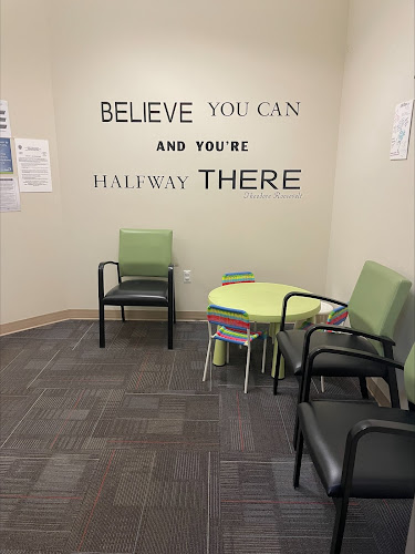 Clinic room with wall text 'BELIEVE YOU CAN AND YOU'RE HALFWAY THERE', seating, and side table with books