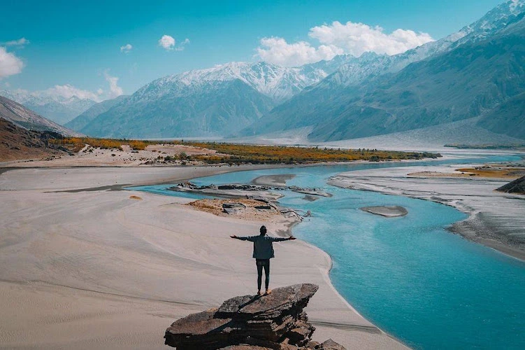 Person standing with arms outstretched on sandy terrain overlooking turquoise river and snow-capped mountain range in Colorado