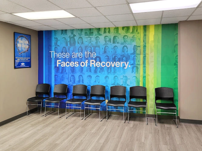Treatment center waiting room with seven black chairs facing a wall mural reading 'These are the Faces of Recovery' with blue and green gradient background featuring portrait photos