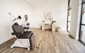 TMS treatment room with black patient chair, white overhead device arm, and clinical equipment