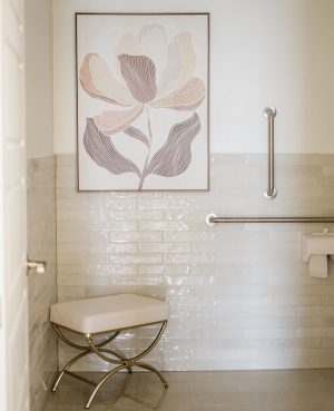 Bathroom with gloss white subway tile, cream bench seat, grab bars, and framed line drawing of flower