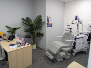TMS treatment room with gray patient chair, purple and white NeuroStar device, wooden desk with supplies, potted plant, and informational poster