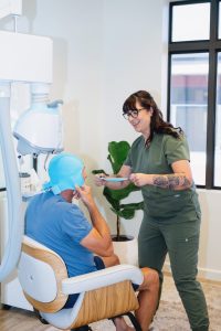 Healthcare provider in green scrubs instructing male patient seated in TMS therapy chair with blue head coil, holding articulated positioning device in bright clinical office
