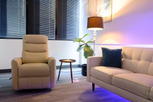 Comfortable waiting area with beige reclining chair, sofa with blue pillow, pendant lamp, and potted plant with purple ambient lighting