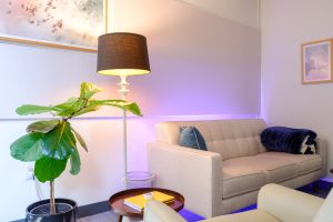 Modern consultation room with cream sofa, potted fiddle leaf fig plant, floor lamp, and purple accent lighting