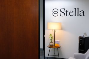 Stella logo on white wall above table lamp and potted plant in reception area