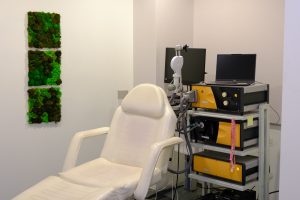 TMS treatment room with white patient chair, medical equipment on cart with monitors, and living moss wall panel