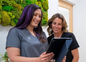 Two women smiling while reviewing information on a tablet in a modern clinic space with moss wall decor