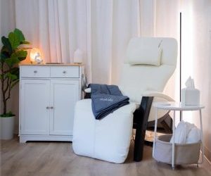 Serene therapy room with white reclining chair, white cabinet with lamp, potted plant, and side table with supplies