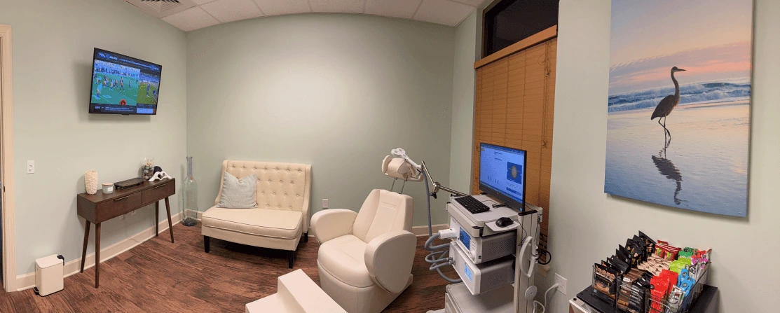 TMS treatment room with beige chair, medical equipment, TV monitors, desk, and heron beach photograph on wall