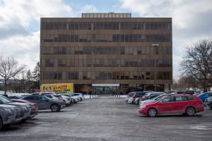 Multi-story brown office building exterior with parked cars and cloudy sky