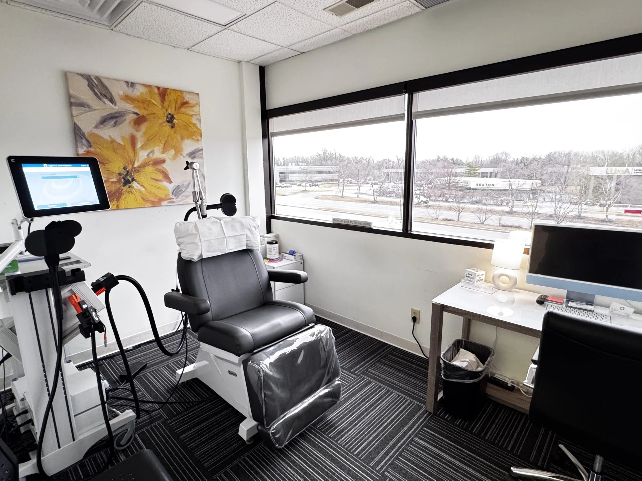 TMS treatment room with patient chair, overhead coil apparatus, monitor, and window view of landscape