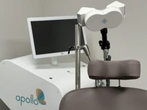 Apollo TMS treatment device with white headpiece mounted above brown patient chair, monitor displaying treatment interface