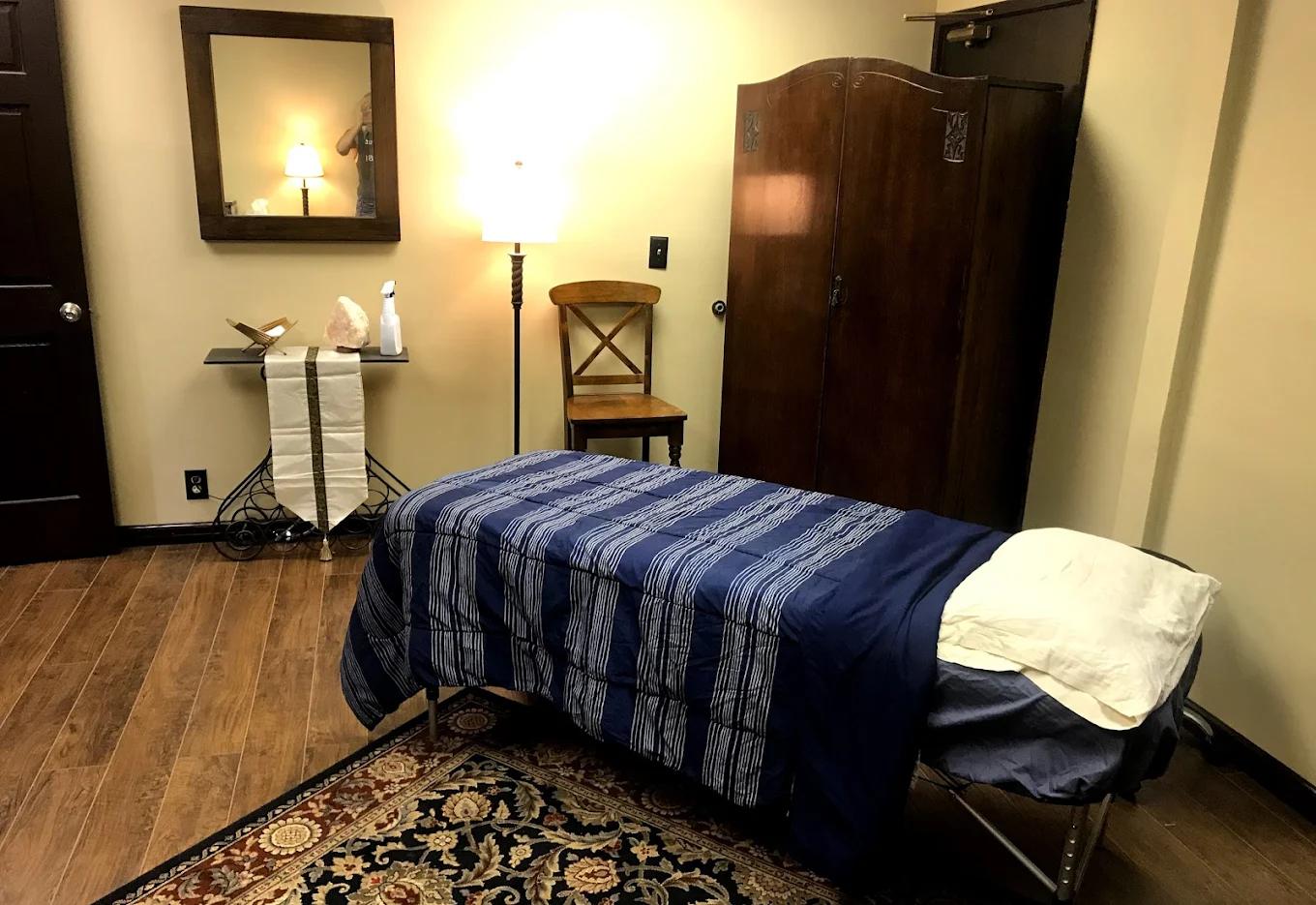 Treatment room with bed, bedside table with lamp, mirror, wooden chair, and dark wood armoire