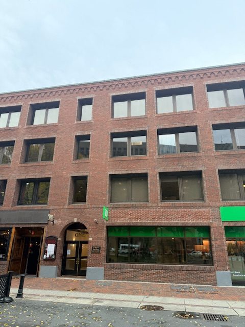 Brick storefront building exterior with large windows and green-lit ground-floor entrance