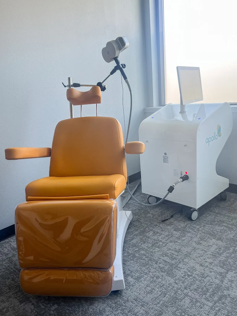 Apollo TMS device next to tan treatment chair with headrest and coil positioning arm in clinical room