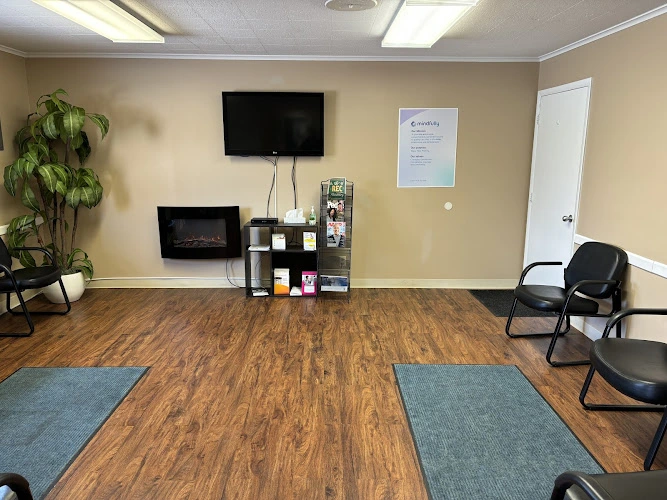Mental health counseling office with beige walls, wooden floor, blue yoga mats, black chairs, potted plant, wall-mounted TV, and fireplace