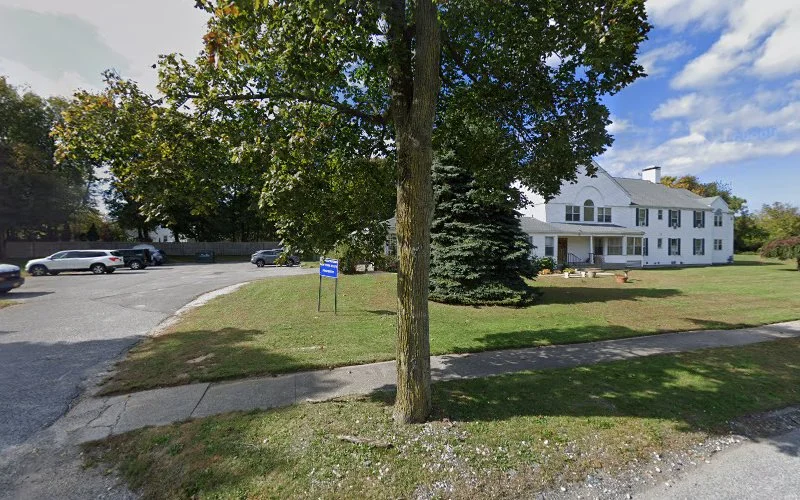 White two-story residential building set on landscaped grounds with large tree and parking area