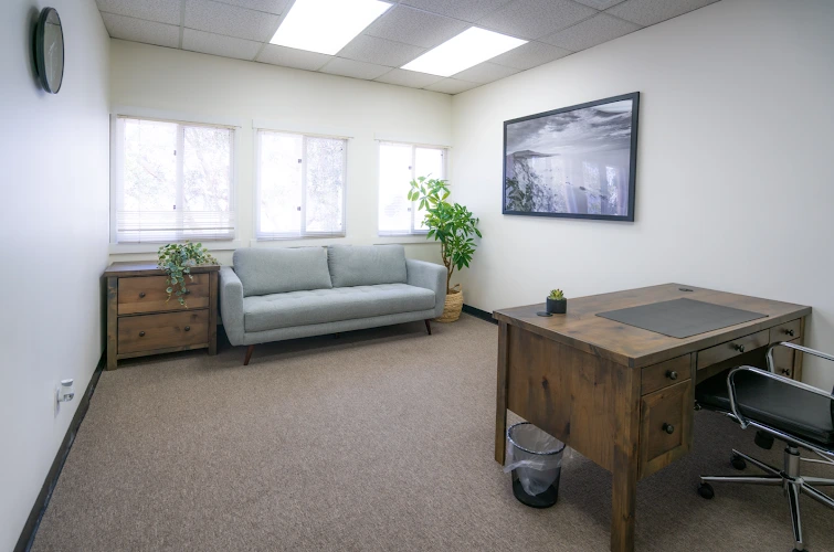 Bright office waiting room with gray sofa, wooden desk, potted plants, large windows, and wall art