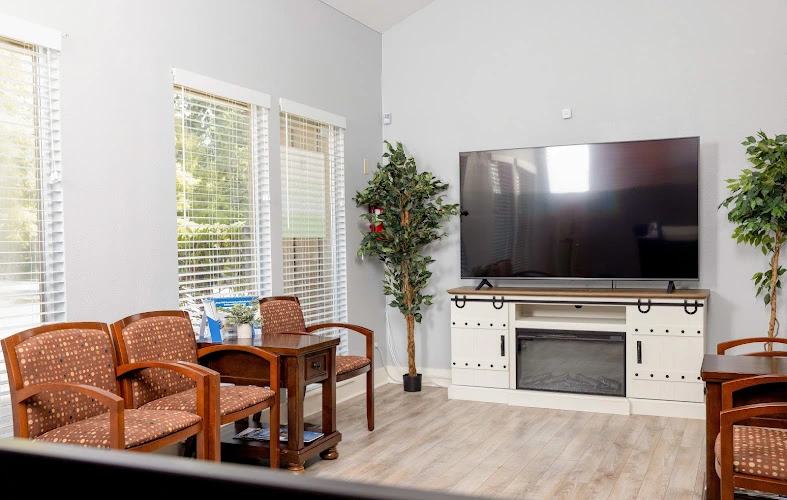Modern clinical waiting room with orange patterned chairs, small table, large wall-mounted TV, potted plants, and bright windows