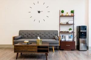 Modern office lounge with gray upholstered sofa, wooden coffee table, wall clock, and shelving unit with plants