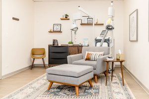 Clinical treatment room with grey armchair, ottoman, desk, shelving, and TMS therapy equipment
