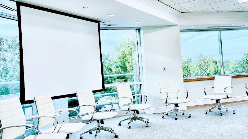 Modern conference room with white chairs, projector screen, and large windows overlooking trees