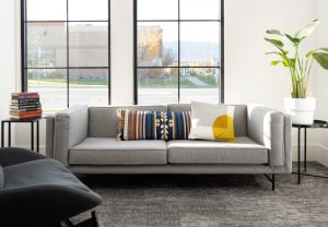 Clinic waiting area with gray upholstered sofa, side tables with books and plants, large windows overlooking grounds