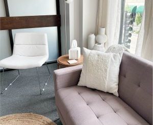 Minimalist clinic interior with purple couch, white chair, and wooden side table near windows