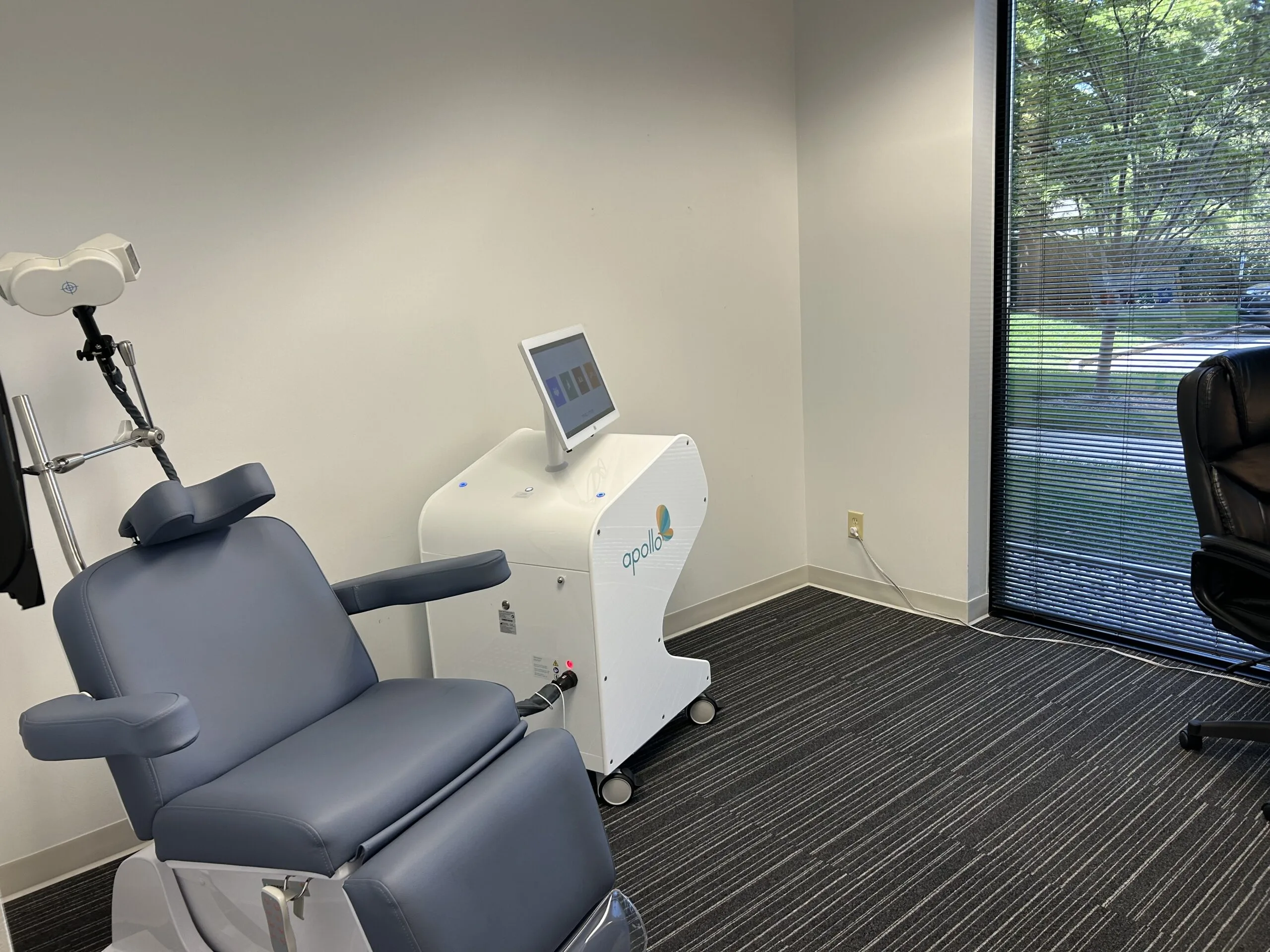 TMS treatment room with patient chair, overhead coil apparatus, and white Oasis TMS device with monitor