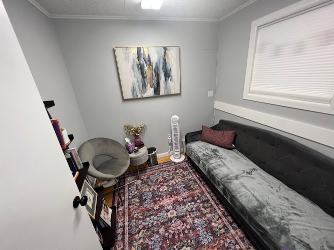 Therapy office with gray walls, abstract artwork, comfortable seating chair, sofa with pillows, burgundy rug, and large window