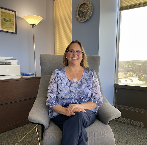 Woman wearing purple patterned top and glasses sitting in gray armchair in office with blue walls