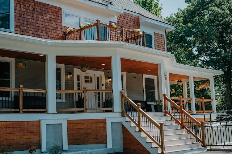 Two-story residential building with brick and blue siding, covered porch, wooden railings, and entrance stairs
