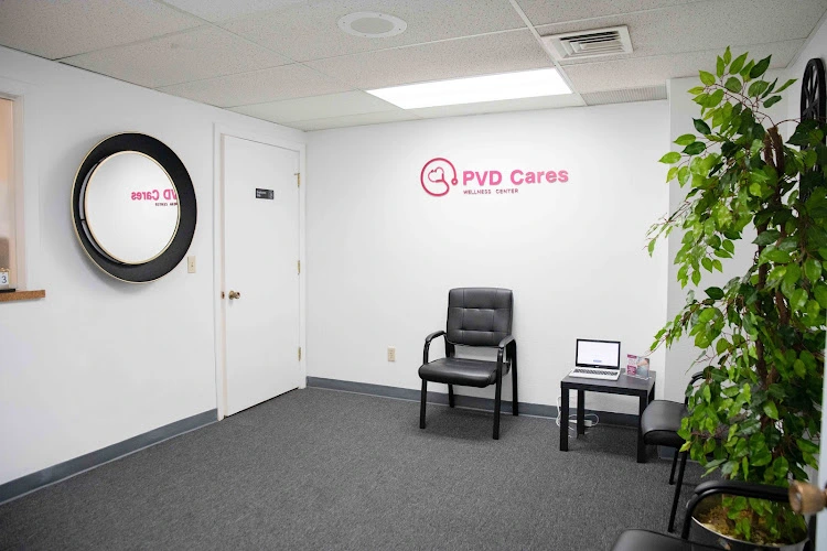 PVD Cares Wellness Center waiting room with pink logo on white wall, black clock, black chair, and green plant