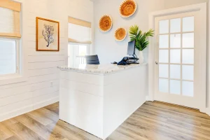 Sweetgrass Psychiatry reception desk with white cabinetry, computer monitor, potted plant, decorative wall art, and natural light from windows