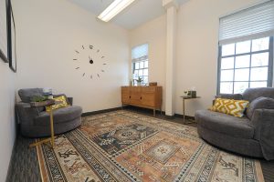 Waiting room with gray upholstered chairs, wooden dresser, geometric patterned rug, and wall clock