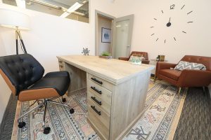 Office space with light wood desk, black and cognac leather chairs, and wall clock