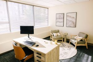 Mental health clinic office with desk, monitor, wooden chair, and seating area with large windows