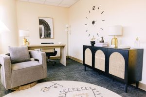 Therapy consultation room with gray armchair, black dresser, wall-mounted clock, and framed artwork