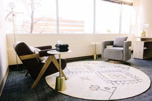 Modern mental health clinic waiting room with black armchair, gray upholstered chair, round patterned rug, and large windows overlooking Denver